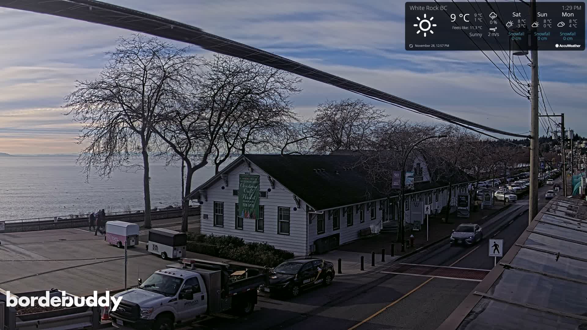 An elevated view captures a sunny 9°C day in a coastal town, featuring a street with vehicles and bare trees beside a white building, all overlooking a calm body of water with distant land.