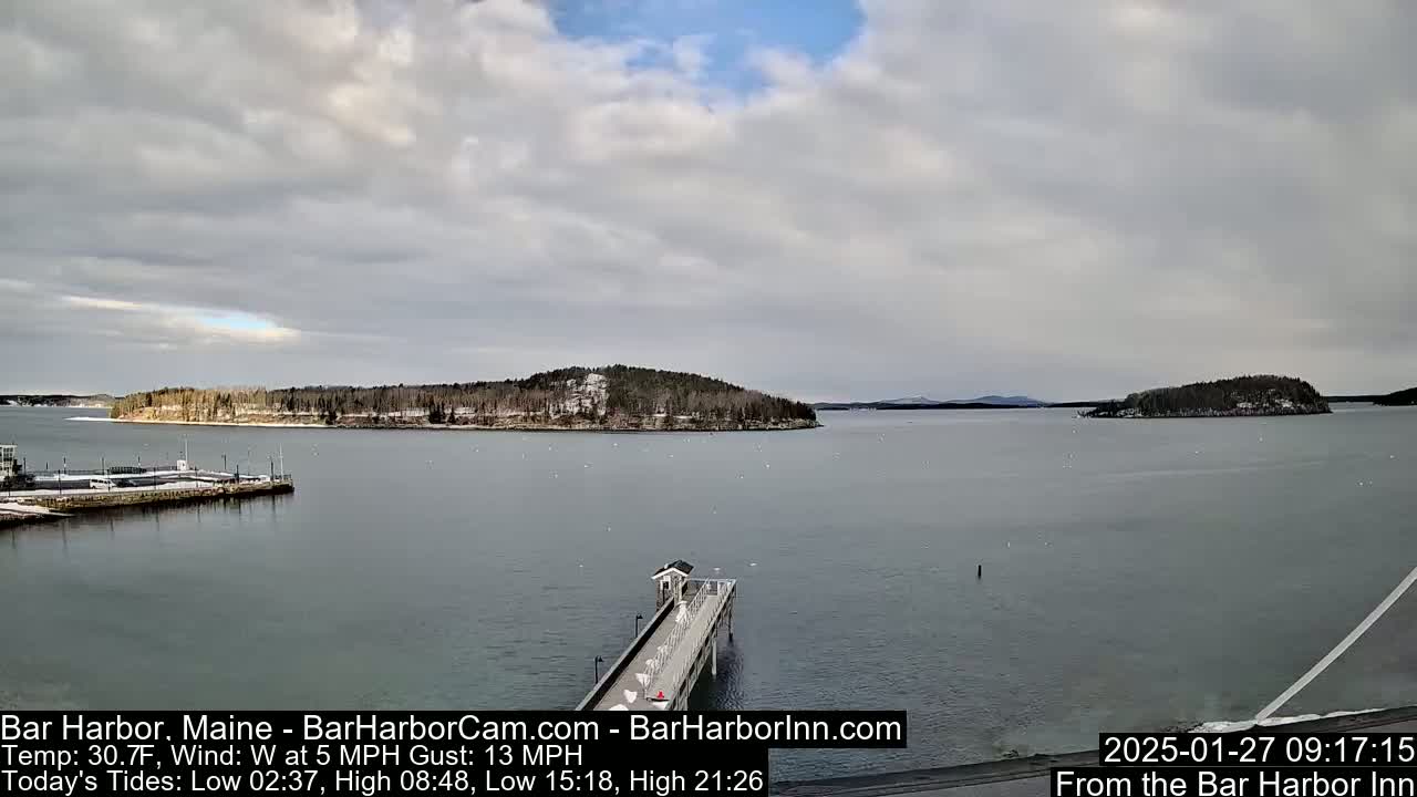 A partly cloudy day shows a snow-dusted island and a long pier extending into a calm body of water.