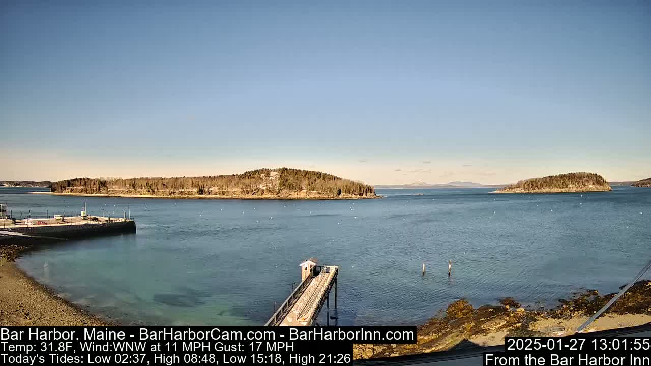 A clear, sunny day shows a calm harbor with a long pier extending into the water, small islands in the distance, and a rocky shoreline.