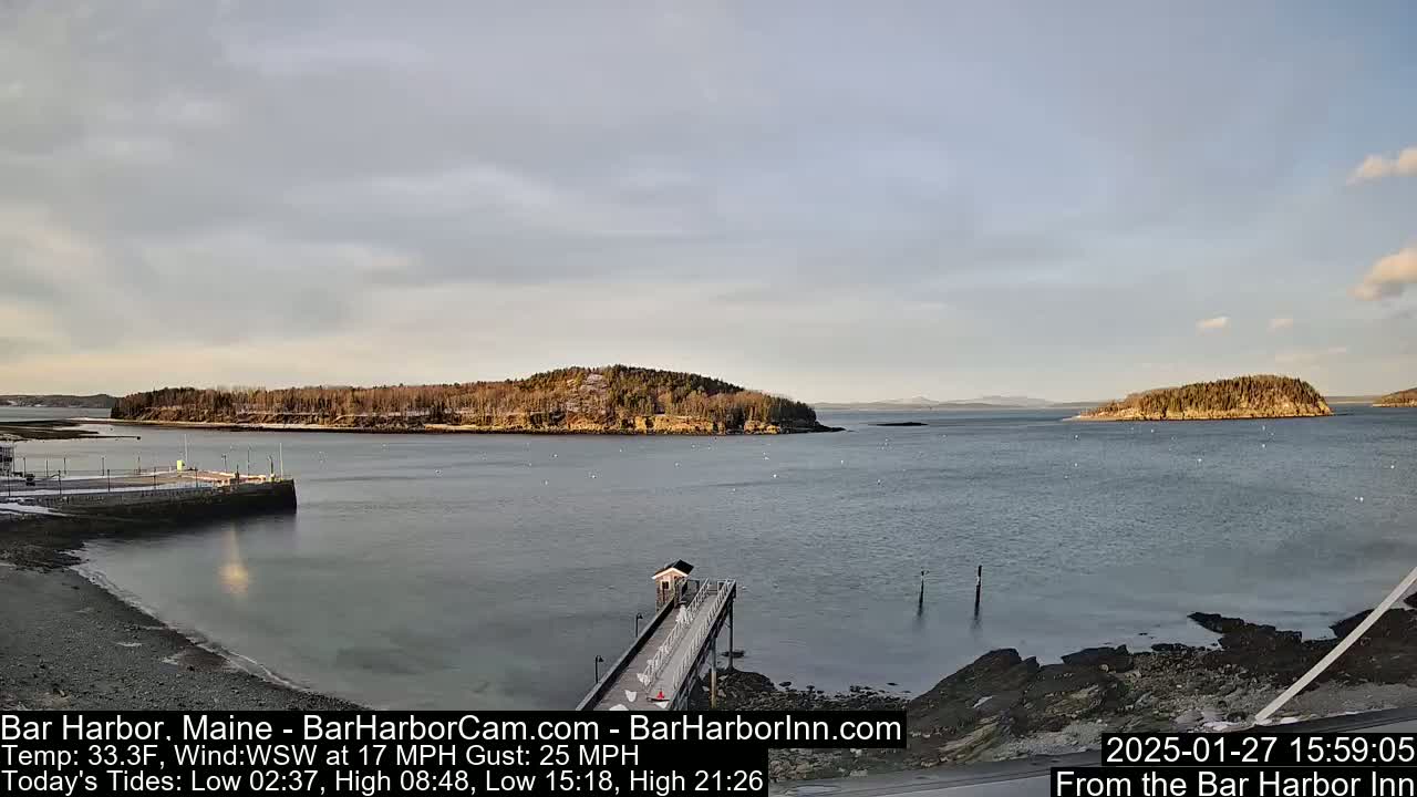 A calm, overcast day shows a view of a harbor with a long pier extending into the water,  two small islands in the distance, and a rocky shoreline.
