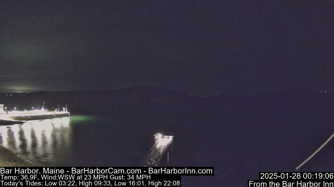 A nighttime view of a calm harbor with a pier extending into the water under a dark sky, with a wind of 23 mph and gusts up to 34 mph.