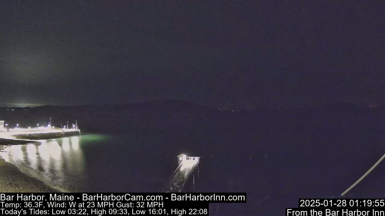 A nighttime view of a calm harbor with a pier extending from a lit shoreline under a dark sky with a 23 mph west wind.
