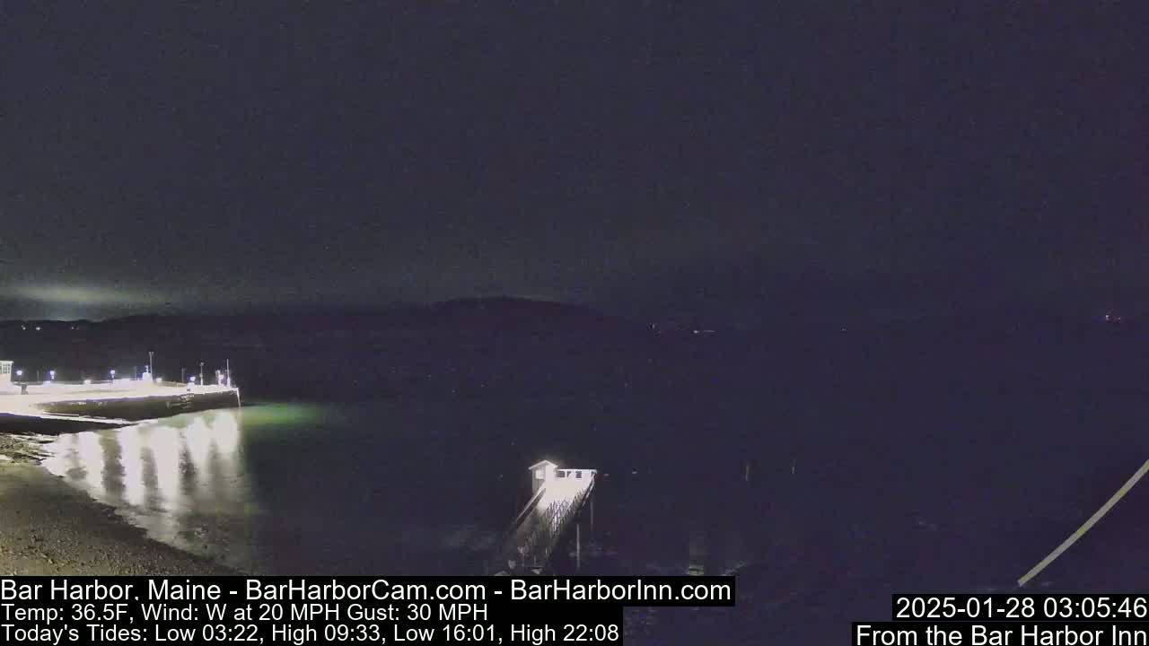 A nighttime, waterfront scene shows a calm body of water reflecting lights from a nearby pier and shoreline under a dark sky with a 20 mph westerly wind and gusts up to 30 mph.