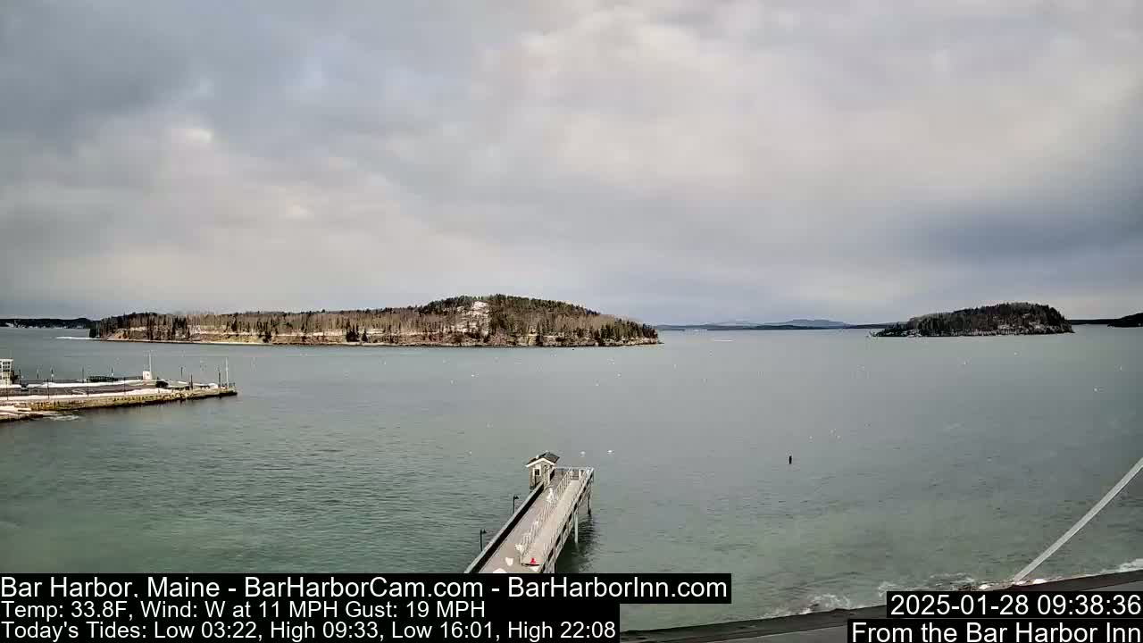 A long pier extends into a calm, gray-sky-covered body of water toward a snow-dusted island, with other islands visible in the distance.