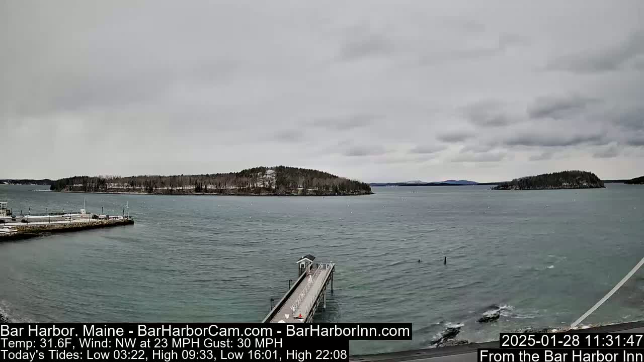 A long pier extends into a calm, gray, overcast ocean toward a snow-dusted island under a cloudy sky.