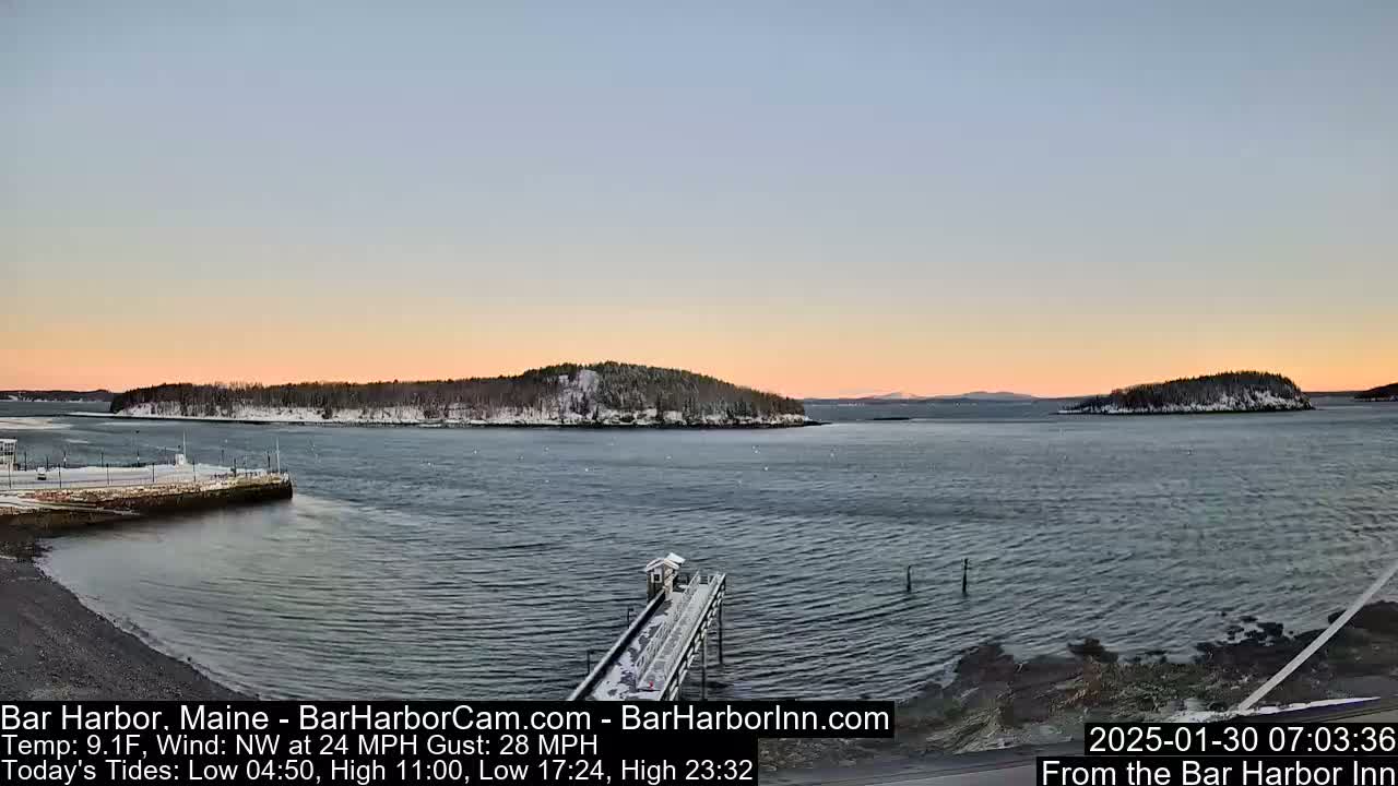 A snow-dusted coastal scene at sunrise shows a calm body of water with a small pier extending from a snow-covered shoreline towards a distant, snow-capped island under a pale pink and blue sky.