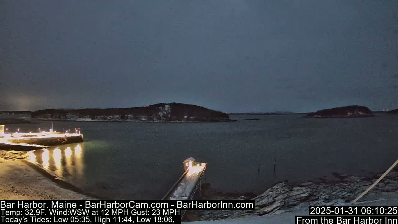 A dark, calm harbor at night is illuminated by lights on a pier and a small building, with a snow-covered island visible in the distance under a twilight sky.