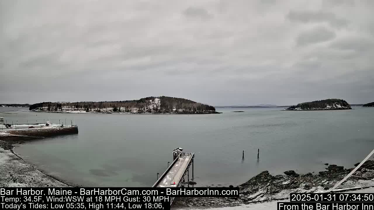 A snow-dusted pier extends into a calm, gray harbor, with several small, snow-covered islands in the distance under an overcast sky.