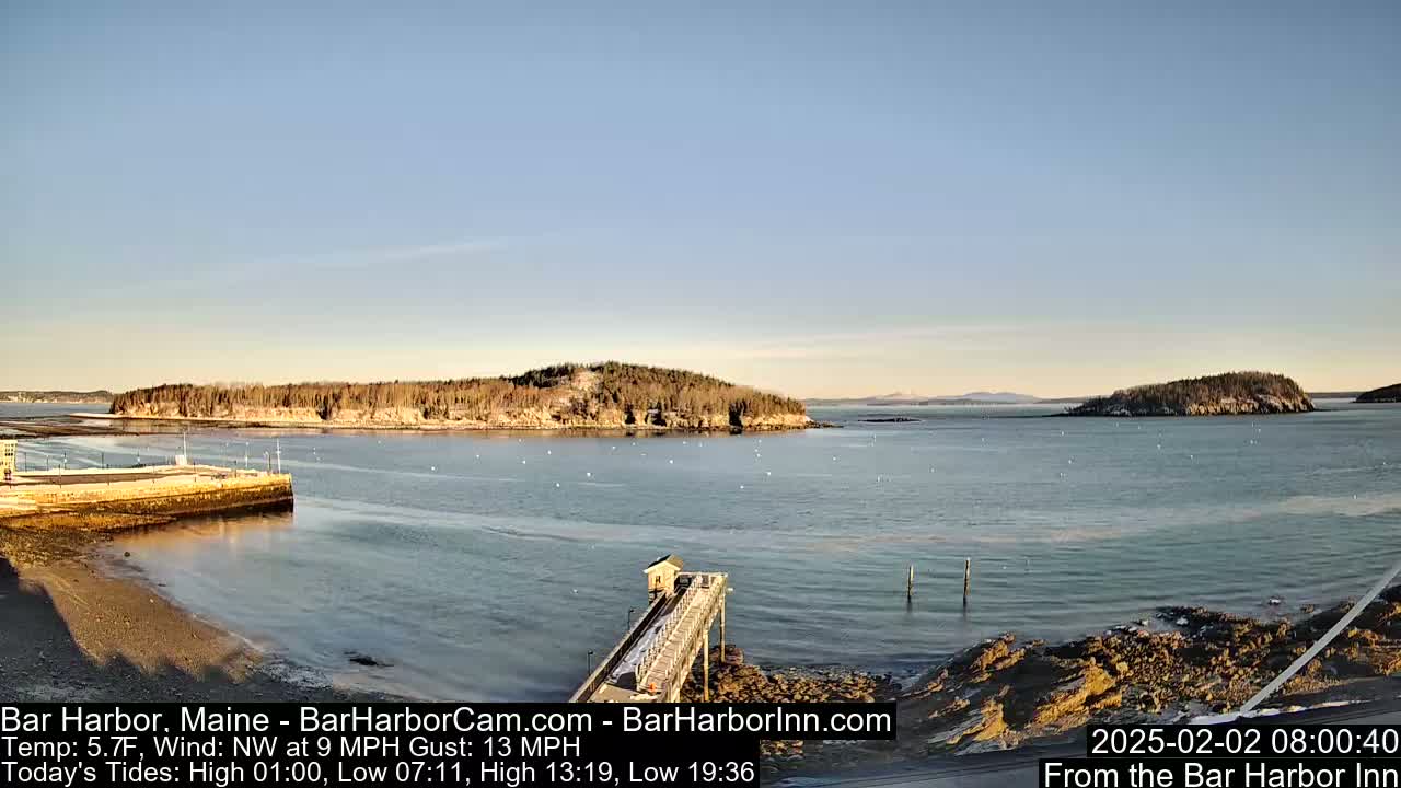 A calm bay with a long pier extending into the water, under a clear, sunny sky, and small islands in the distance.