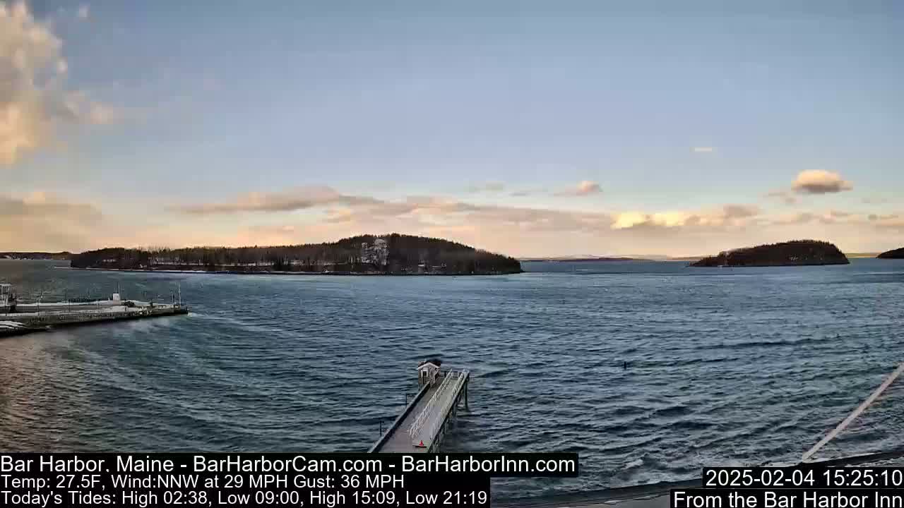 A partly cloudy, windy day overlooks a choppy body of water with a long pier extending from a snow-dusted shoreline to the ocean, featuring several small islands in the distance.