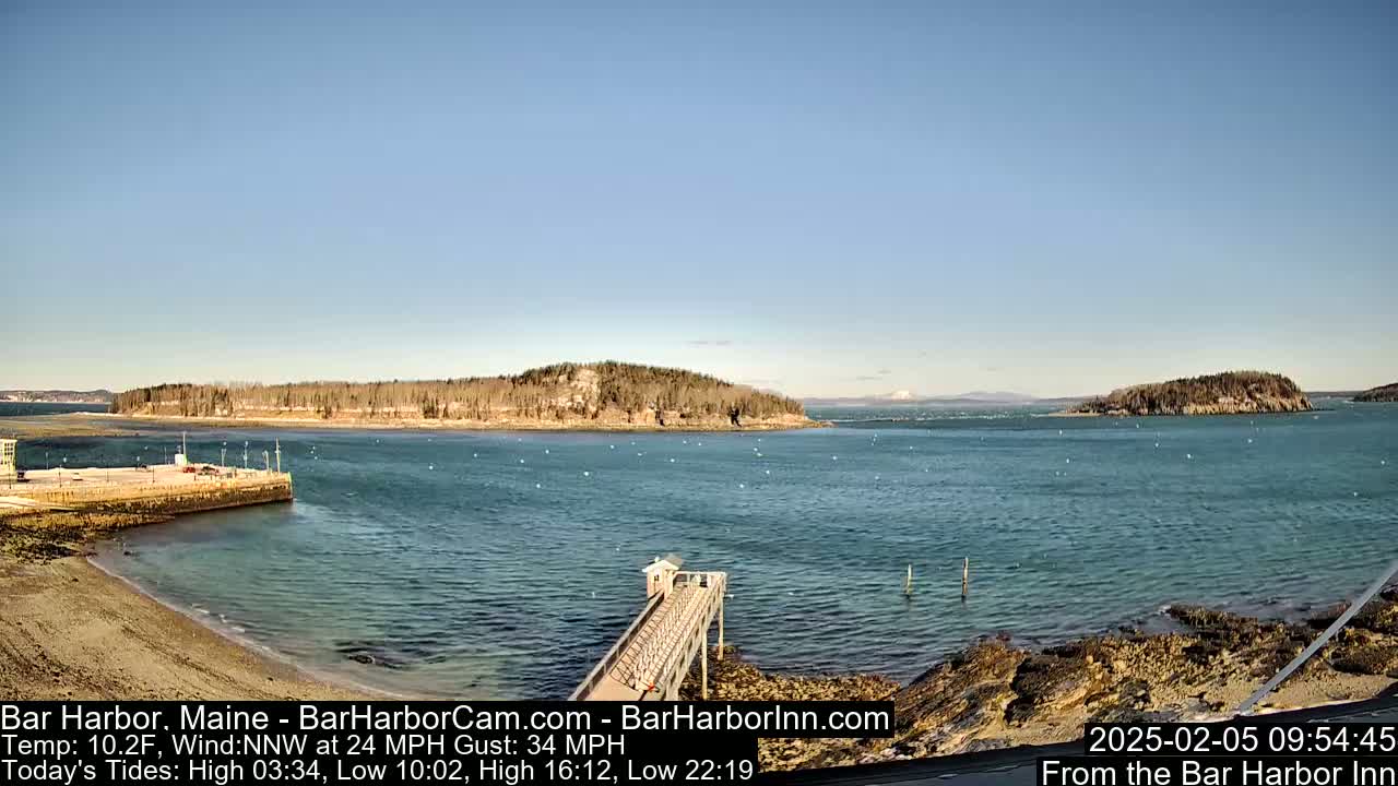 A calm, blue harbor with a long pier extending from a sandy shore into the water, featuring several small islands in the distance under a clear sky.