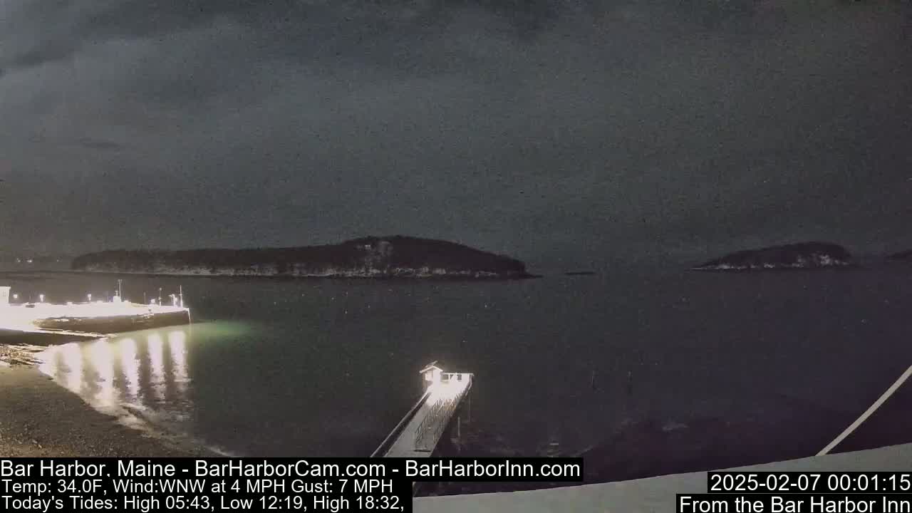A nighttime, snowy scene shows a dark body of water with a lit-up pier, and small islands in the distance under a dark, cloudy sky.