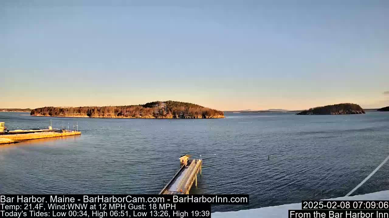 A calm body of water with islands in the distance under a clear, pale blue sky is viewed from a pier.