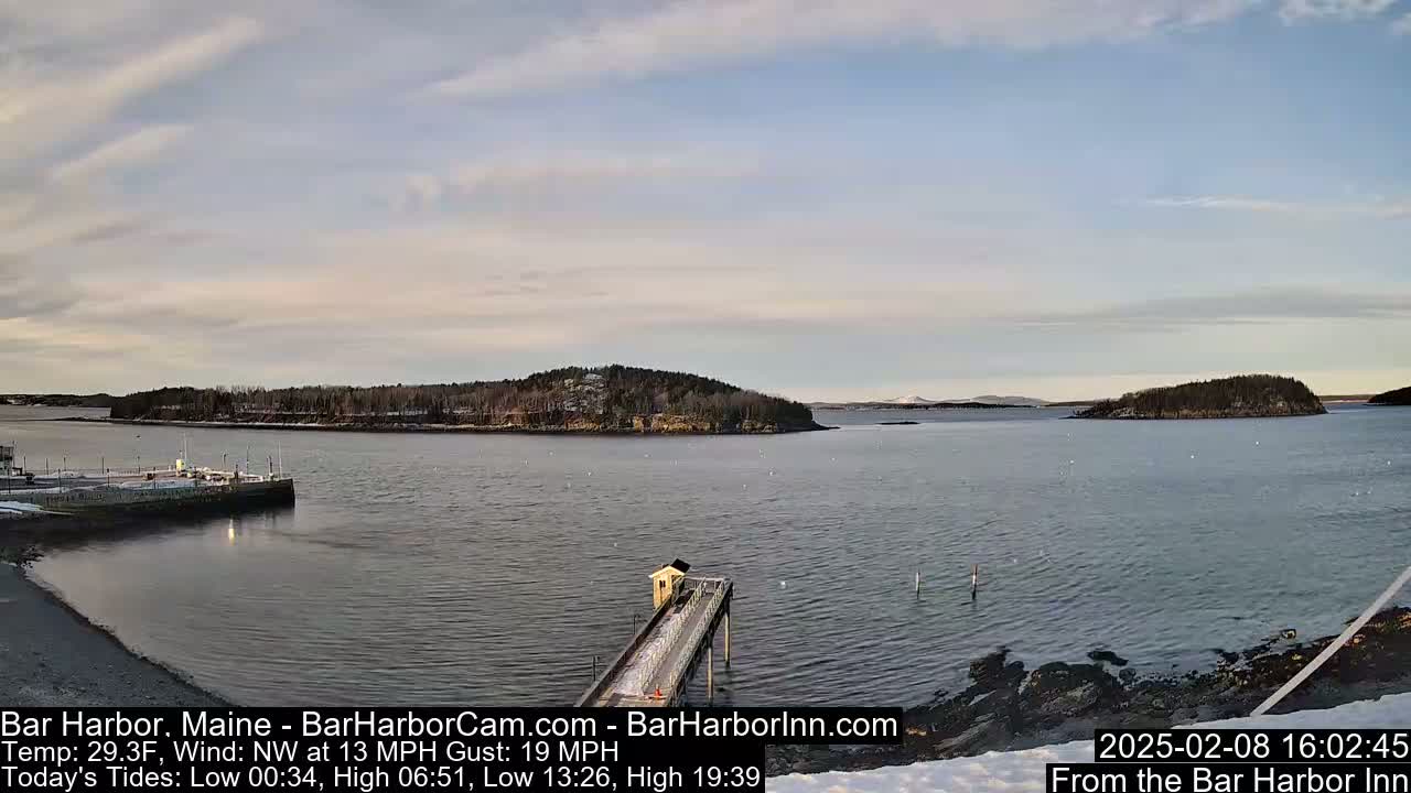 A partly cloudy day shows a calm body of water with small islands and a pier extending from a snowy shoreline.