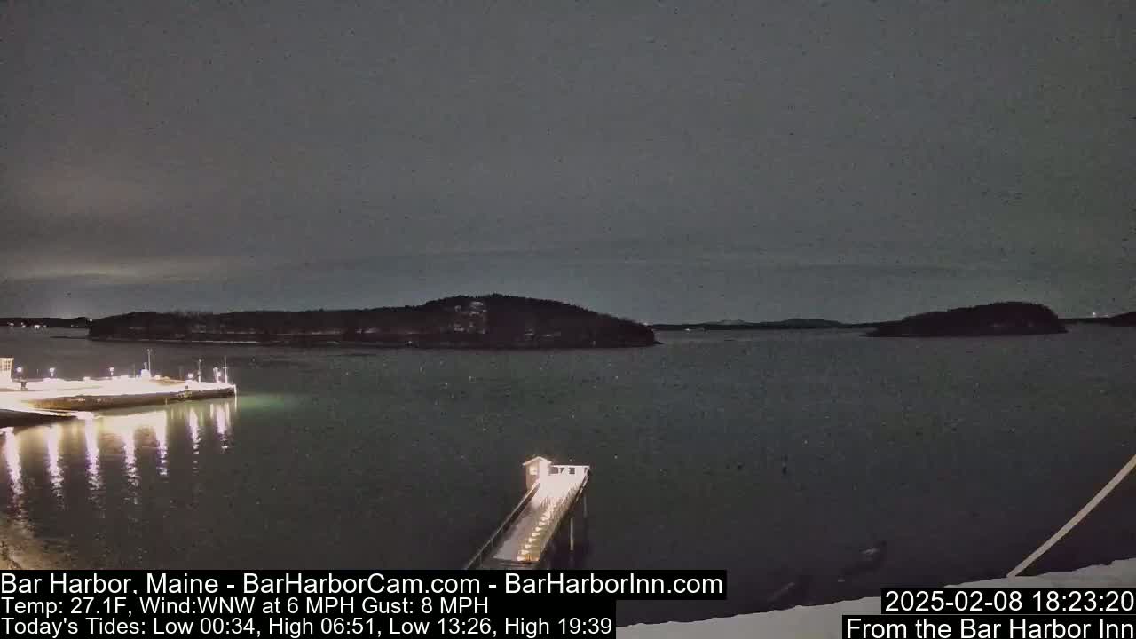 A nighttime, snow-dusted scene shows a dark body of water with illuminated piers and distant islands under a gray sky.