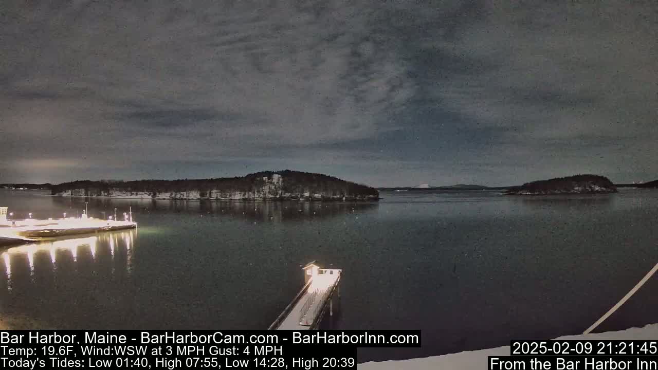 A nighttime view of a calm, dark body of water with snow-covered islands and a lit pier extending from the shore under a cloudy sky.