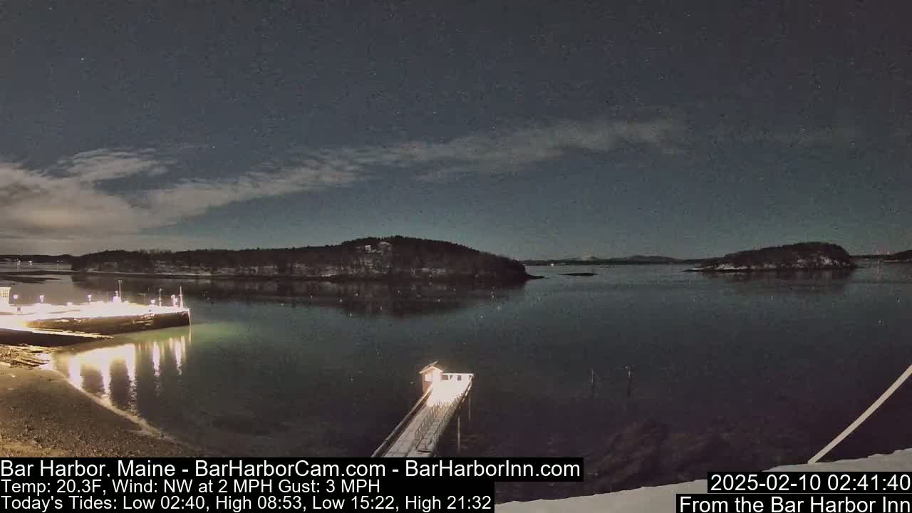 A nighttime view of a calm harbor under a partly cloudy sky dotted with stars, showing a lit-up pier and a snow-covered shoreline.