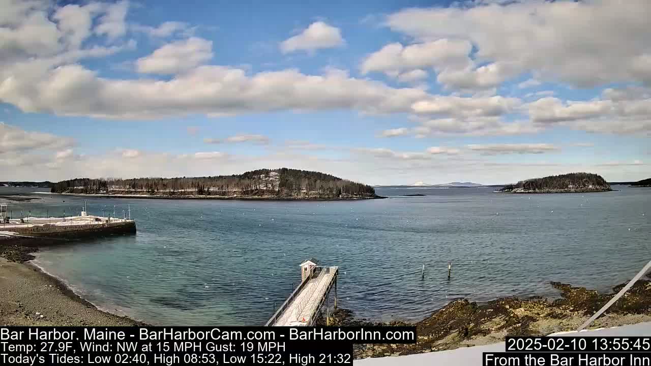 A partly cloudy day reveals a rocky coastline with a long pier extending into a calm body of water, featuring several islands in the distance, some with snow cover.