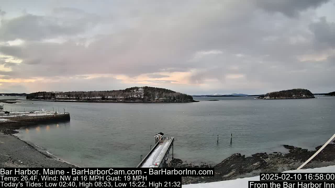 A snow-dusted harbor scene under a cloudy sky shows a pier extending into calm water, with islands in the distance.