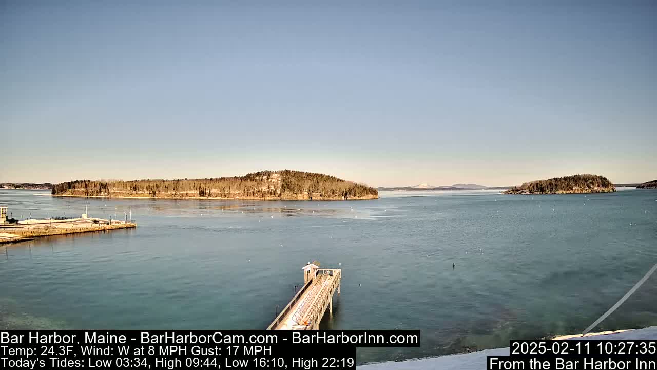 A long pier extends into a calm, blue body of water, with snow-covered islands visible in the distance under a clear, light blue sky.