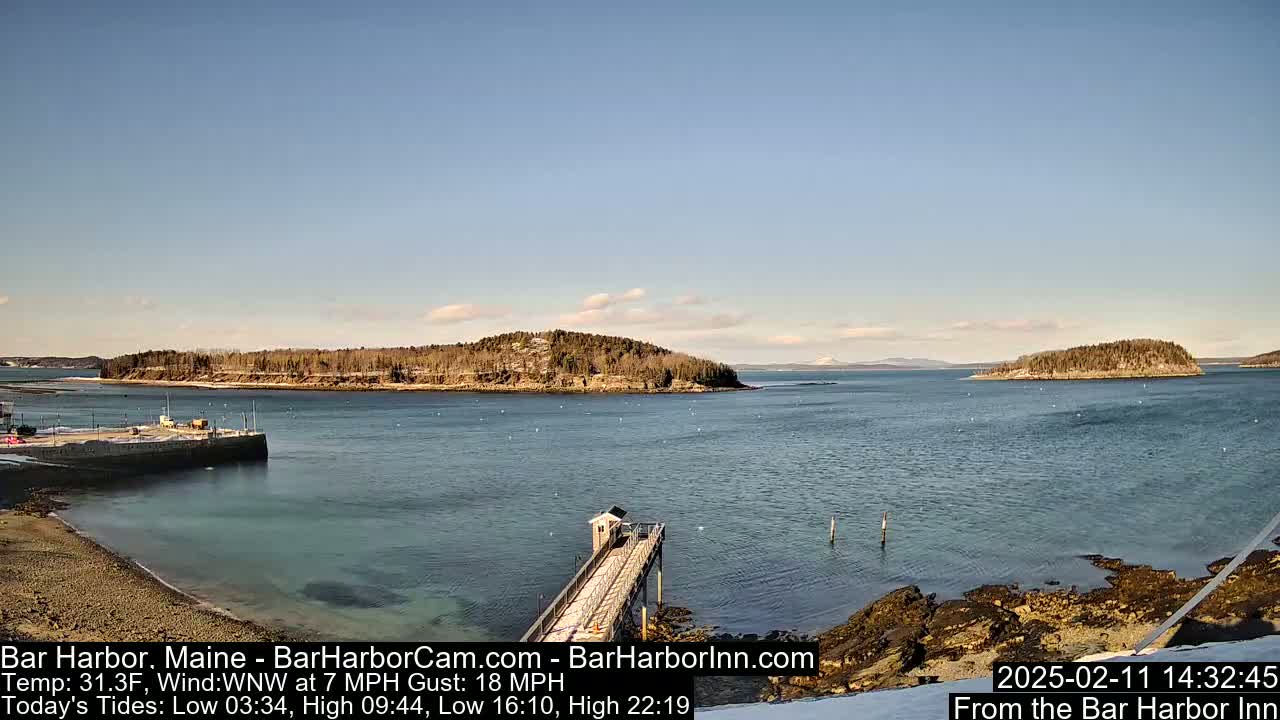 A calm, partly sunny day reveals a harbor with a long pier extending into the water, several small islands in the distance, and a shoreline with rocky outcrops.