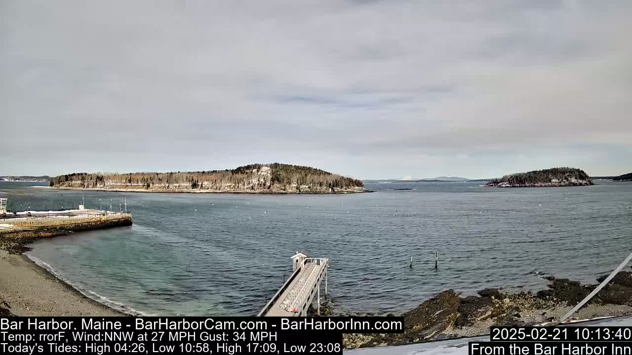 A tranquil harbor scene on a cloudy day shows a long pier extending into the water, with several small islands in the distance, some showing patches of snow.