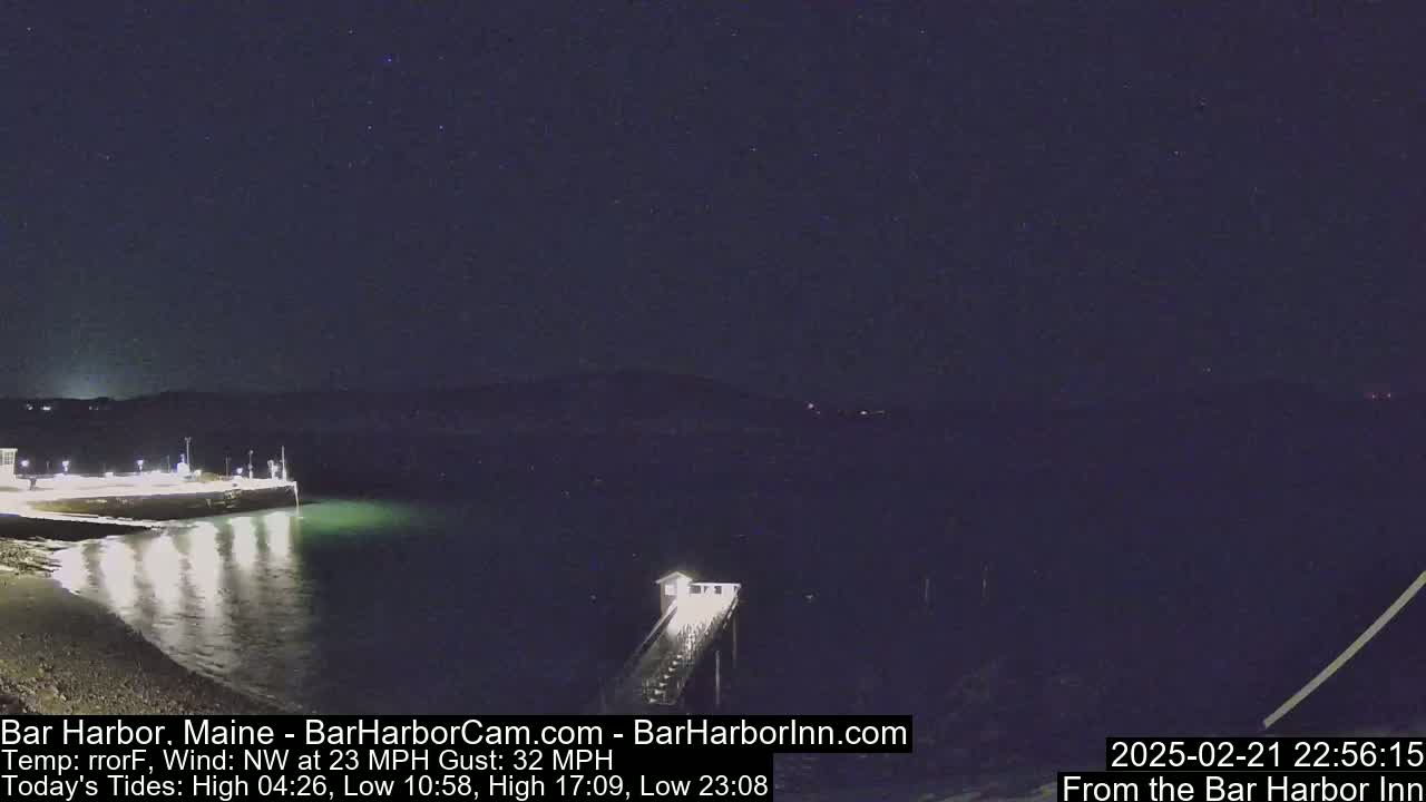 A nighttime view of a calm body of water with a pier extending from a lit shoreline under a starry sky with a northwest wind at 23 mph and gusts up to 32 mph.