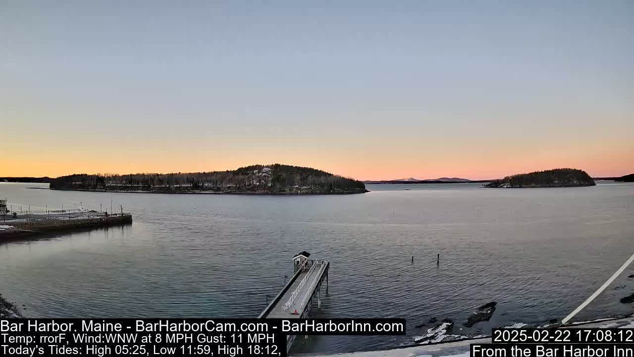 A calm body of water at sunset, with a long pier extending from a snow-dusted shoreline toward a small island, under a pale pink and orange sky.