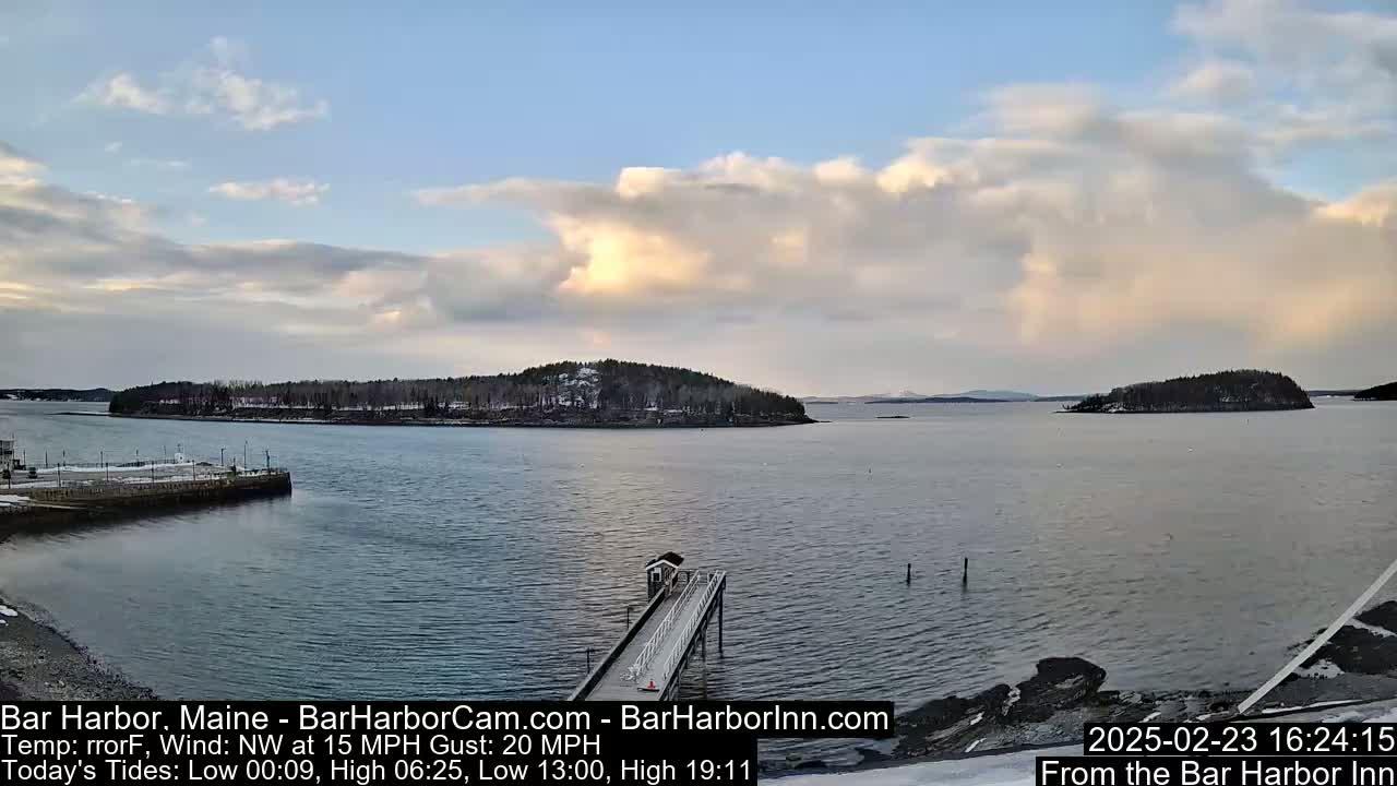 A partly cloudy, breezy day overlooks a calm harbor with a long pier extending into the water, several small islands, and snow-covered shoreline.