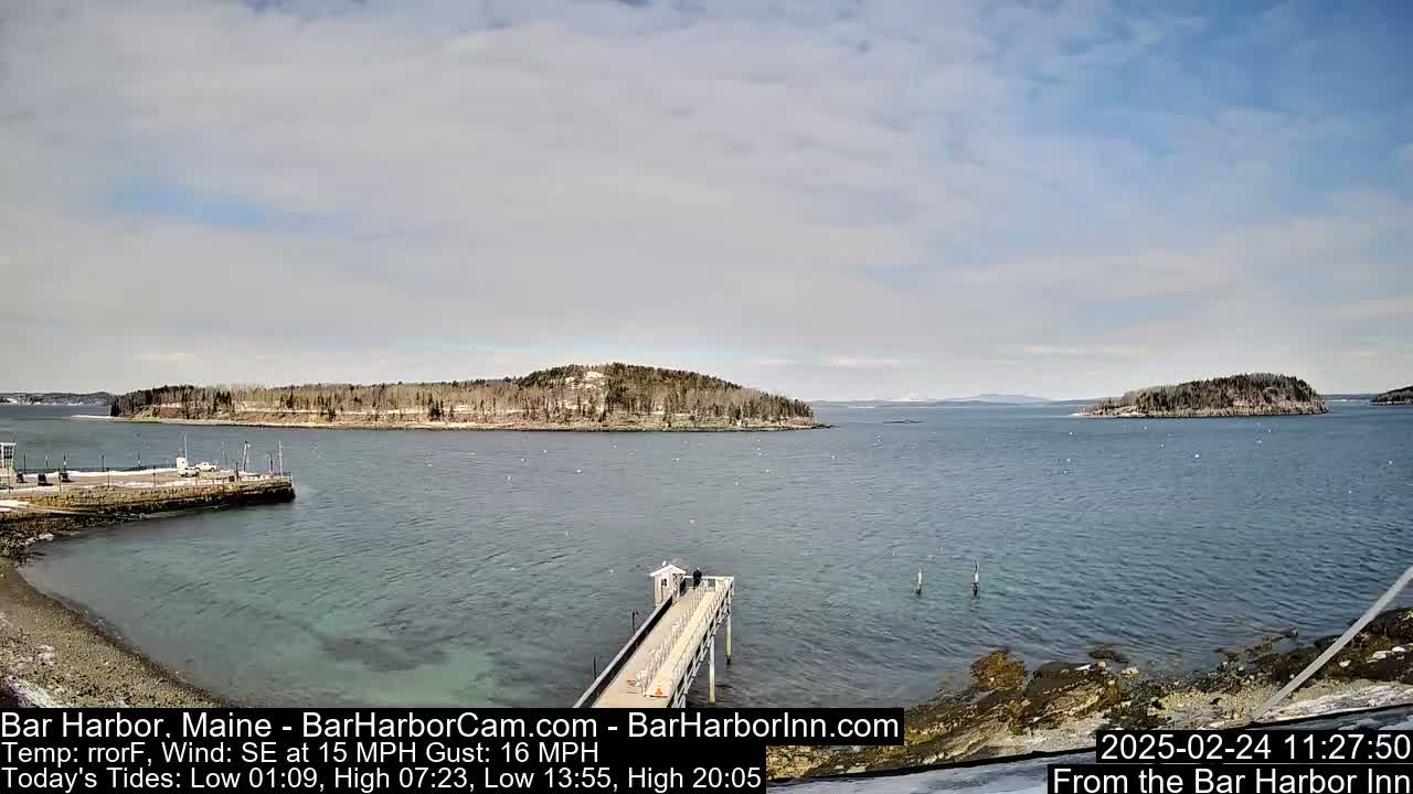 A long pier extends over calm, clear water toward two small, tree-covered islands under a partly cloudy sky.