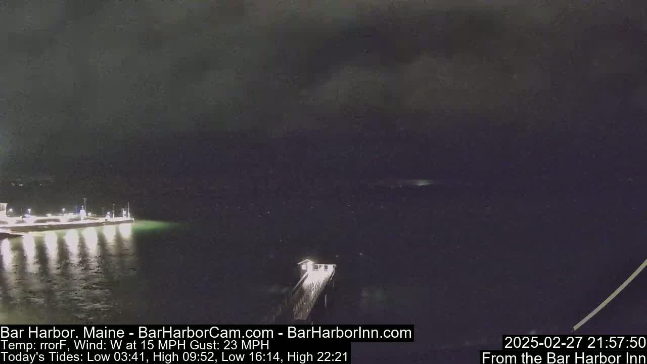 A nighttime view of a dark body of water with a pier and lit shoreline under a cloudy sky with a 15 mph wind and 23 mph gusts.
