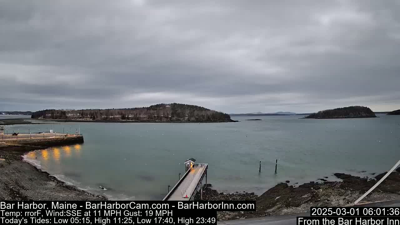 A mostly cloudy day reveals a calm bay with a long pier extending from a rocky shoreline toward a small, tree-covered island, with another larger landmass in the distance.