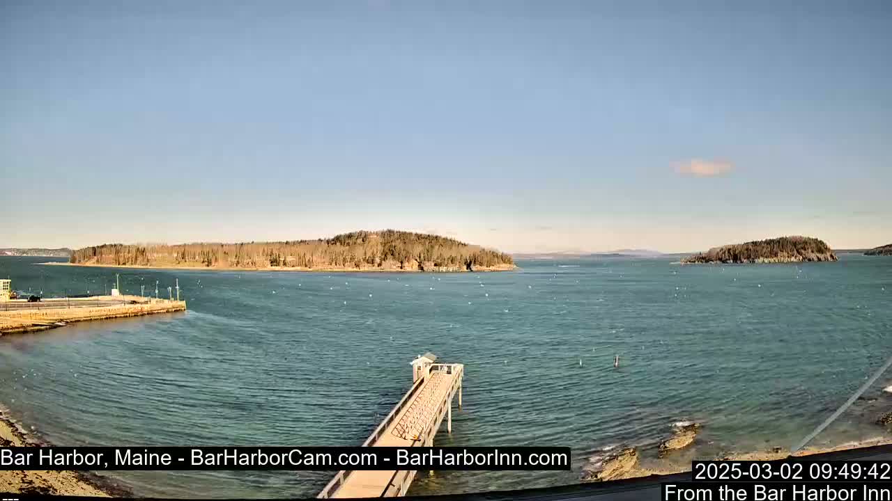 A tranquil harbor scene on a sunny day shows a long pier extending into the water, with several small islands visible in the distance.