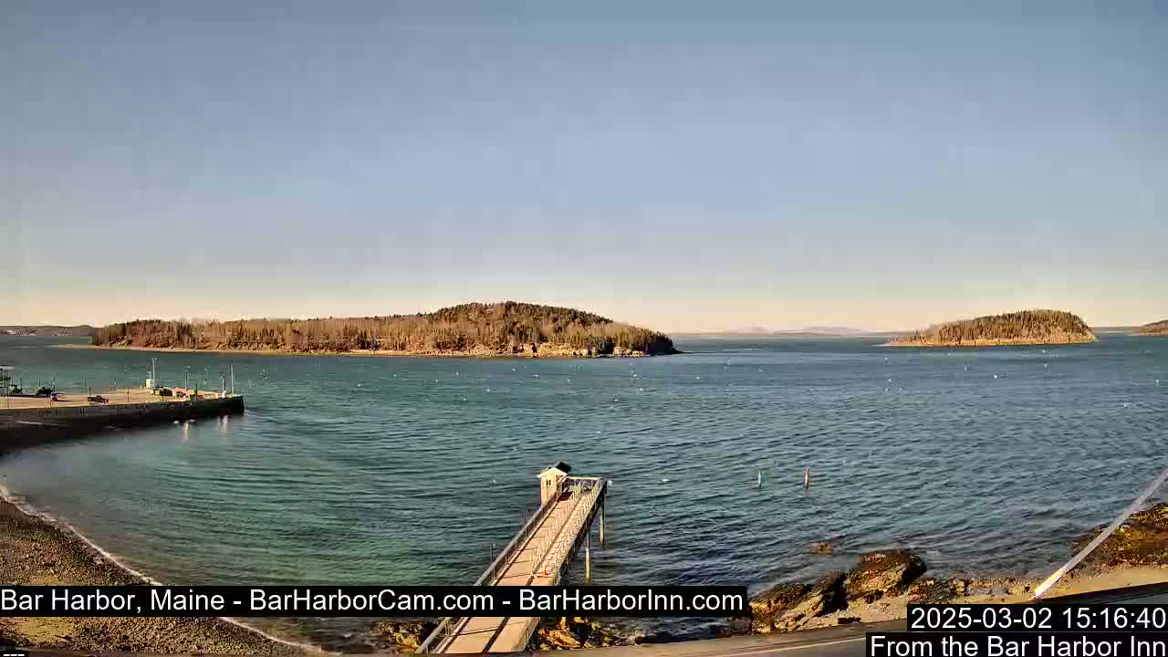A calm, sunny day reveals a harbor scene with a long pier extending into the water, small islands in the distance, and a shoreline with rocky areas.