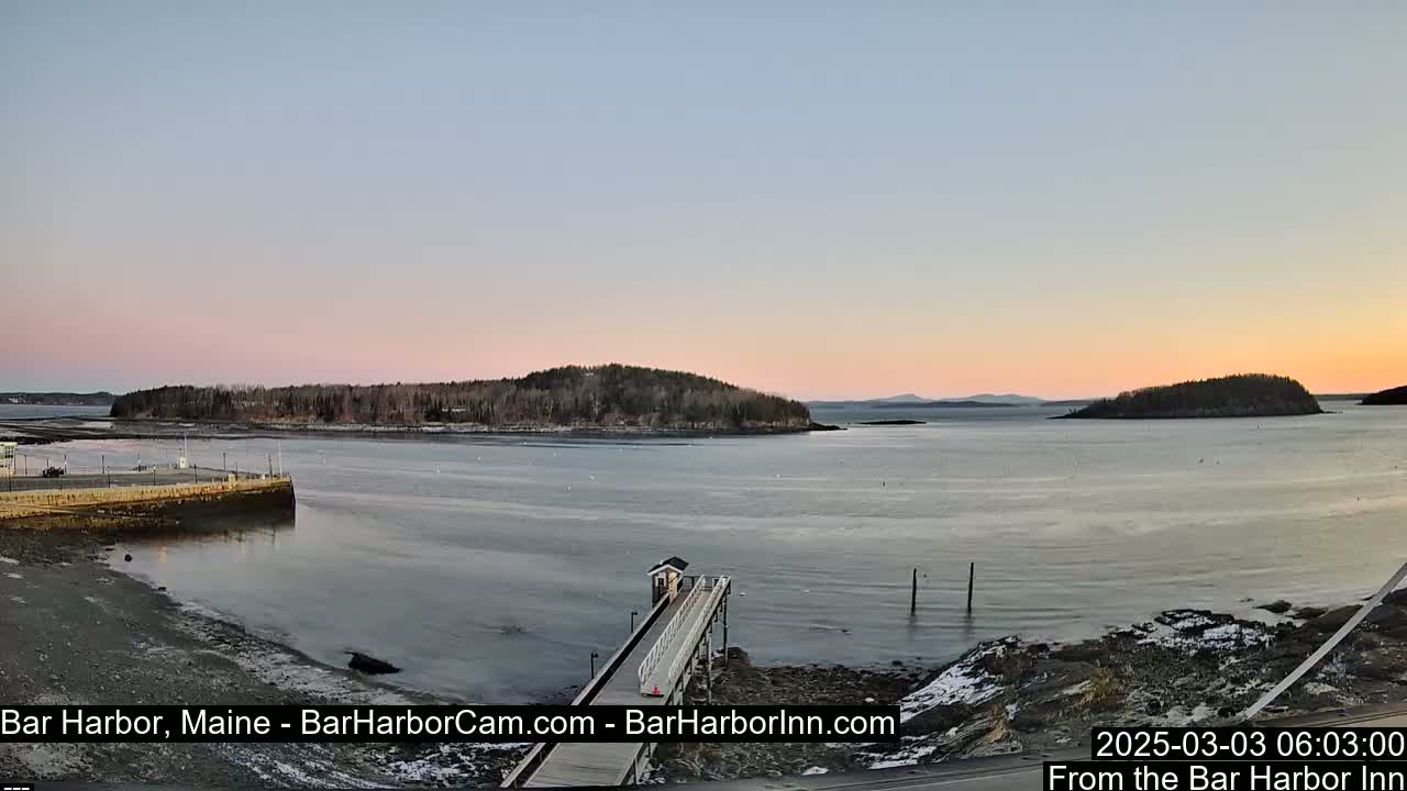 A tranquil harbor scene at sunrise shows a calm body of water partially frozen, with a small pier extending from the shoreline toward a small island in the distance, under a pale pink and blue sky.