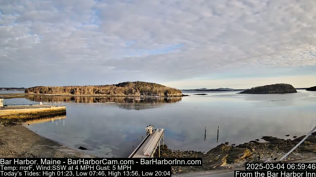 A calm body of water with a small island in the distance under a partly cloudy sky is seen from a high vantage point, showing a long pier extending into the water from a rocky shoreline.