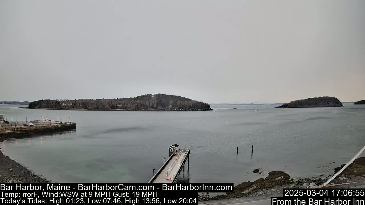 A calm body of water, under an overcast sky, features a long pier extending from a rocky shoreline toward a small island in the distance.