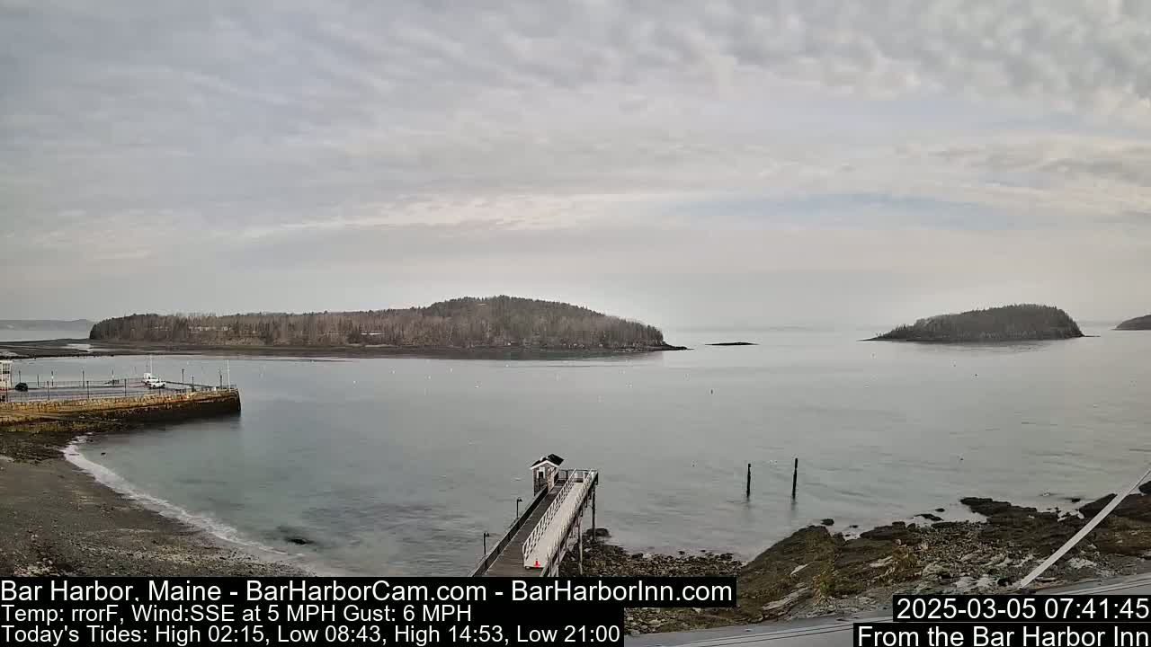 A calm, overcast day reveals a rocky shoreline with a pier extending into a bay, and several small, tree-covered islands in the distance.