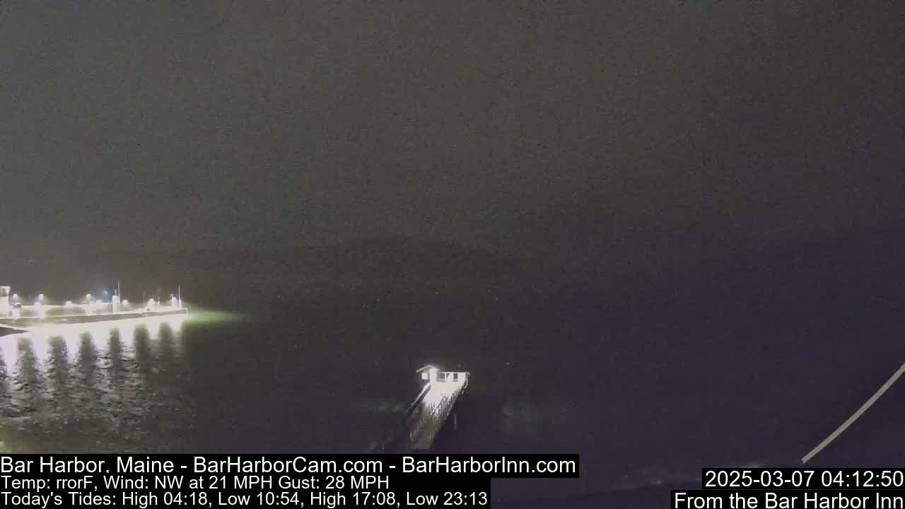 A nighttime, dimly lit view of a pier extending into dark water under a cloudy sky with a strong northwest wind.