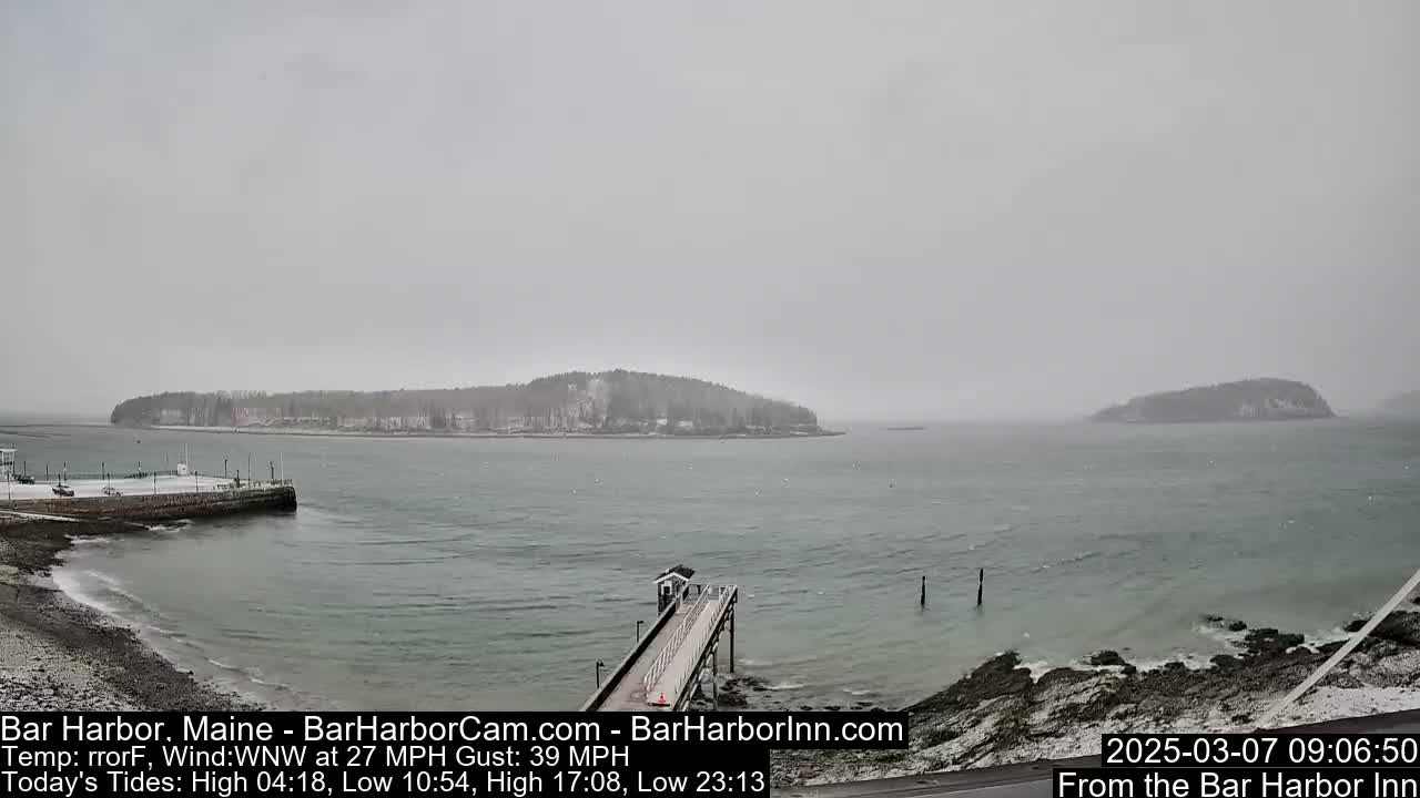 A snow-covered pier extends into a gray, choppy ocean toward two snow-dusted islands under an overcast sky.
