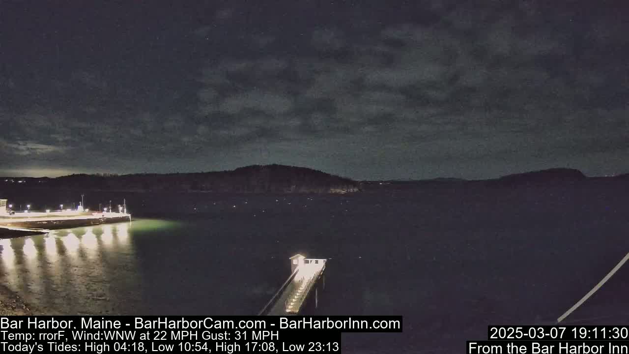 A nighttime coastal scene shows a dark body of water under a mostly cloudy sky, with a lit pier extending from a shoreline into the water, and lights illuminating a nearby area.