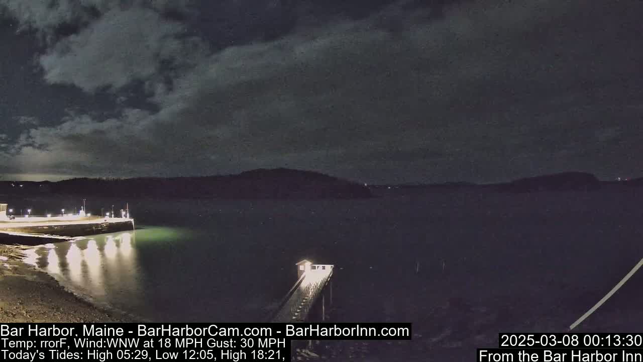 A nighttime view of a calm body of water with a lit pier extending from a shoreline under a partly cloudy sky.