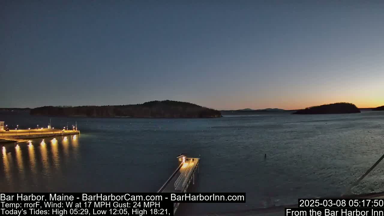 A twilight view of a calm body of water, with a lit pier extending from a shoreline into the water, and islands in the distance under a clear sky.