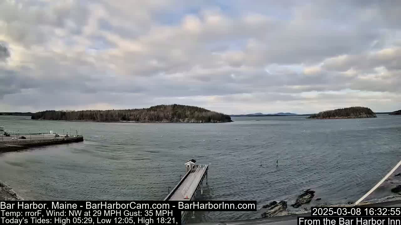 A mostly cloudy day shows a calm body of water with a long pier extending from a rocky shoreline, and islands in the distance under a mostly cloudy sky.