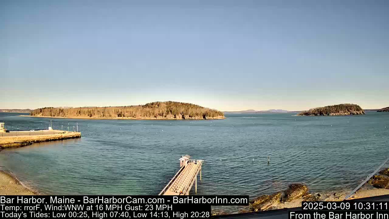 A calm, clear day shows a body of water with islands in the distance, a long pier extending into the water, and a rocky shoreline in the foreground.