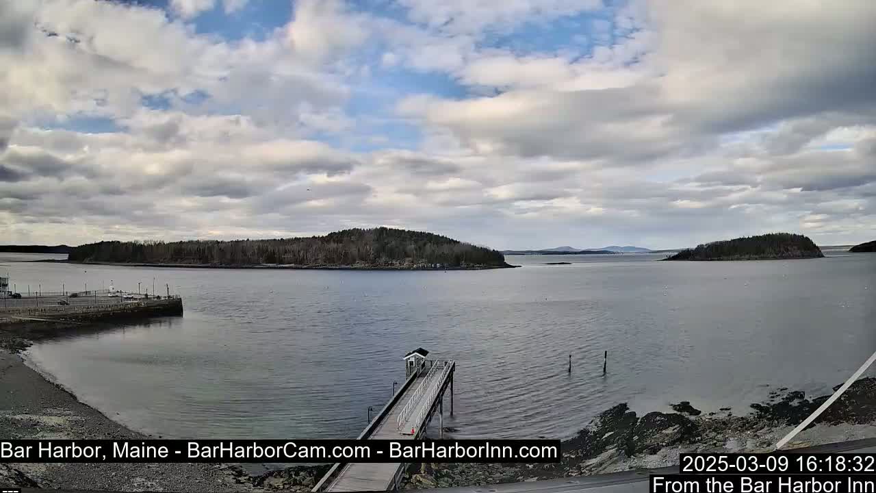 A partly cloudy day reveals a tranquil harbor scene with a long pier extending into the water, tree-covered islands in the distance, and a shoreline with a parking area.