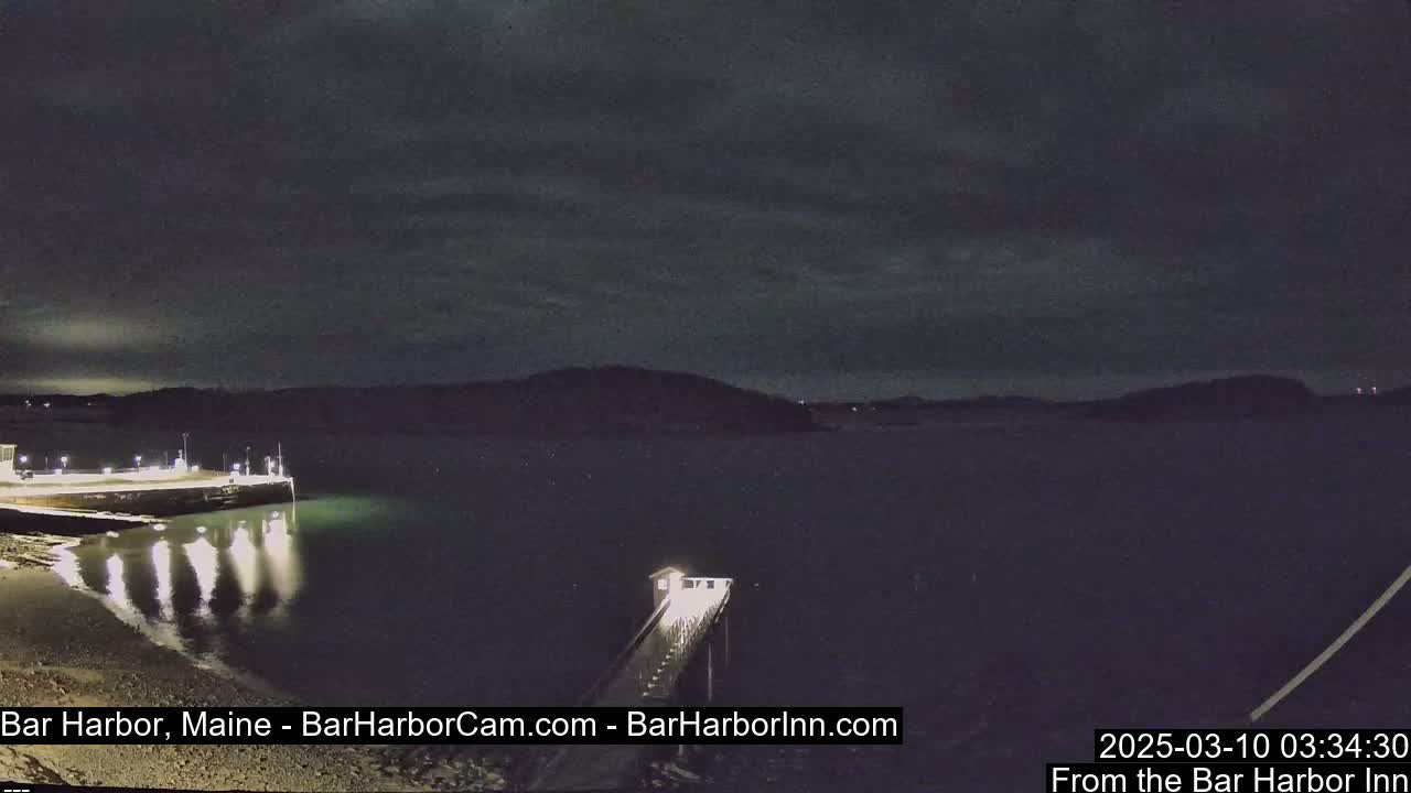A nighttime view of a dark, calm harbor with a lit pier extending into the water under a mostly cloudy sky.