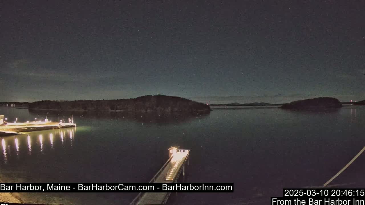 A nighttime view of a calm body of water with a lit pier extending from a shoreline, across which a small island is visible under a clear, dark sky.