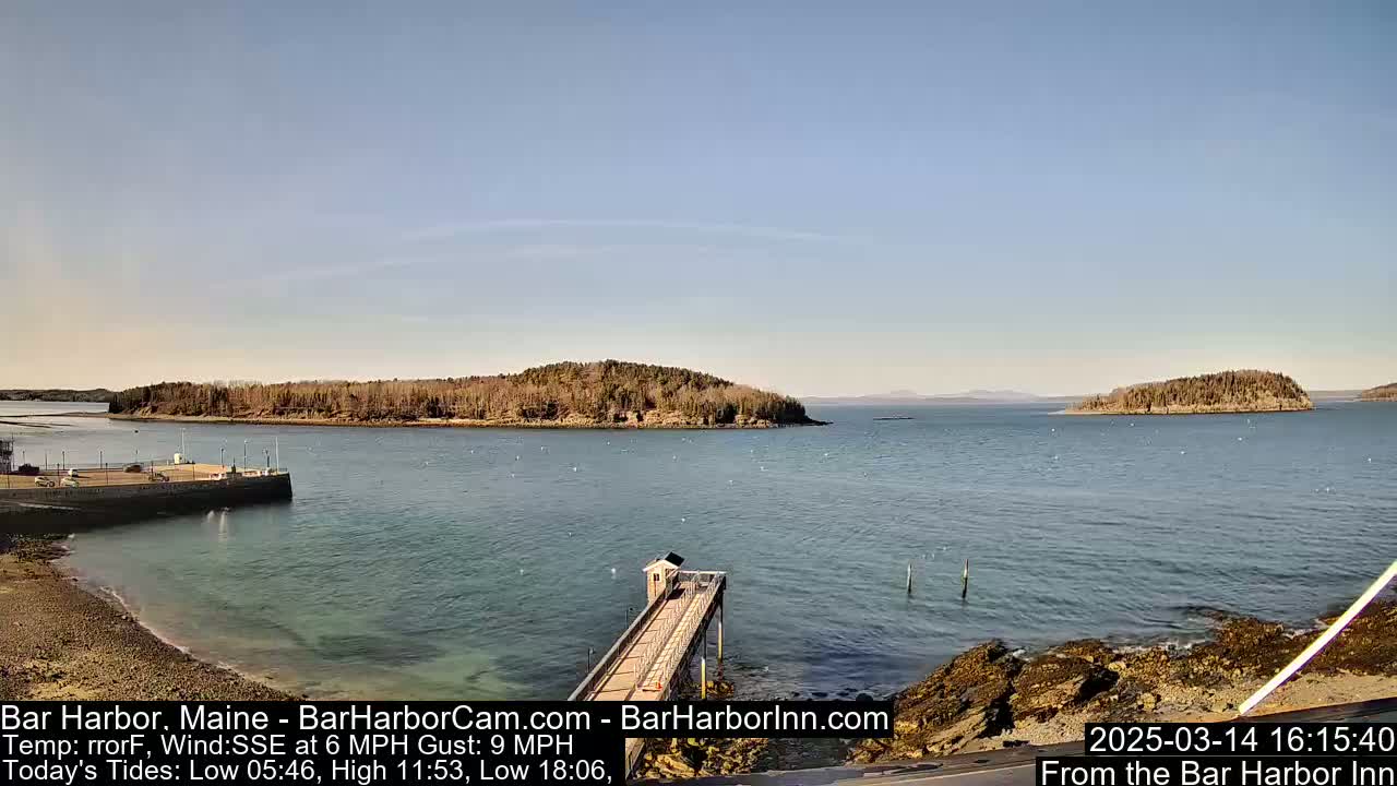 A tranquil harbor scene on a sunny day shows a pier extending into calm water, with several small islands visible in the distance.