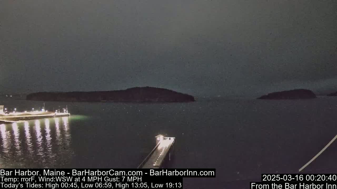 A dark, nighttime scene shows a calm body of water with a lit pier extending from a shoreline toward a small, distant island under a cloudy sky.